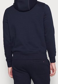 Sudadera de color navy con capucha, con dobladillo y puños acanalados, hecha de un tejido suave. La vista trasera muestra un diseño minimalista y detalles de costura.