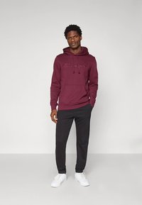 Bordo jopa s kapuco s logotipom »Champion«, kombiniran s črnimi ohlapnimi trenirkami in belimi supergami, stoji na nevtralnem ozadju.