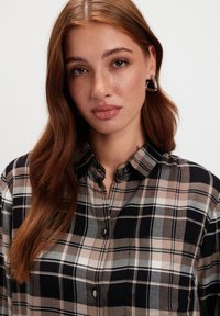 Młoda kobieta z długimi rudymi włosami, nosząca srebrne trójkątne kolczyki oraz koszulę w kratę w kolorach czarnym, beżowym i białym.