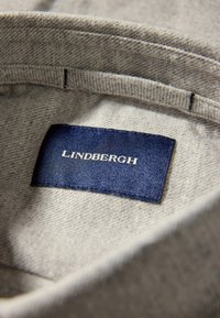 Grijze stof met een textuurpatroon, met een blauwe label waarop "LINDBERGH" in witte tekst staat, bevestigd aan de binnennaad van een kledingstuk.