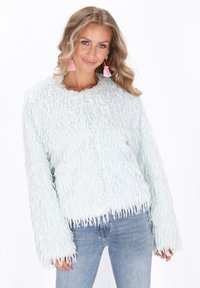 Frau in einem hellblauen, strickigen Sweater und hellblauen Jeans, die mit rosa Quasten-Ohrringen lächelt, vor einem weißen Hintergrund.