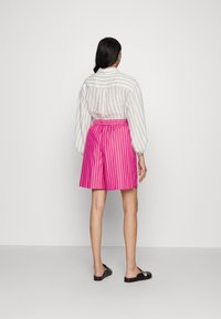 Bílá pruhovaná košile s dlouhým rukávem je kombinována s růžovými šortkami s vertikálními pruhy. Šortky mají elastický pas a končí nad koleny. Černé slip-on boty.