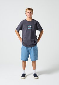 Grafisches T-Shirt aus dunkelgrauer Baumwolle mit Frontdruck. Kombiniert mit hellblauen Jeansshorts. Das Model trägt weiße Socken und marineblaue Schuhe.