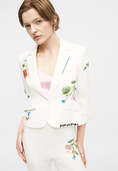 Γυναίκα που φοράει λευκό ραμμένο σακάκι και παντελόνι με πολύχρωμη κεντητή λουλουδένια διακόσμηση και λεπτομέρειες από στρας, συνδυασμένα με ανοιχτό ροζ μπλουζάκι.