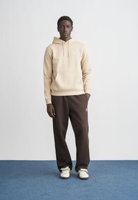 Béžová mikina s kapucí a přední kapsou, kombinovaná s tmavě hnědými tepláky a bílými teniskami. Jednoduchý, volný střih s logem jako doplňkem.