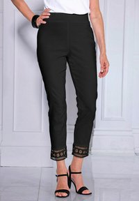 Zwarte tapercut broek heeft een gestructureerde zoom met uitsneden, gecombineerd met zwarte sandalen met bandjes. De outfit heeft een gladde afwerking en minimale details.