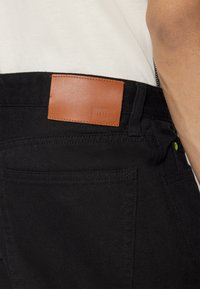 Sorte denimbukser med en glat struktur, der har en brun læderpatch med teksten "HUF" ved taljen og grønne syninger.