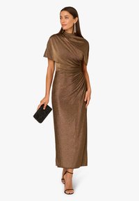 Abito lungo metallico marrone con design drappeggiato e maniche a cappotto corte. La modella tiene una pochette nera con texture e indossa sandali neri con fascia.