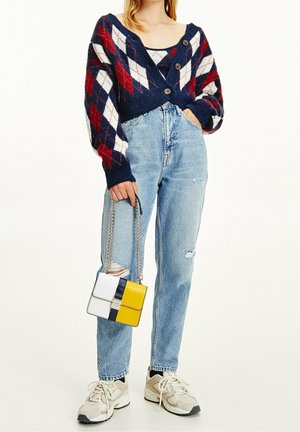 Woman wearing blue, red, and white argyle cardigan and tank top, light ripped jeans, white sneakers, holding a white and yellow handbag with chain strap.