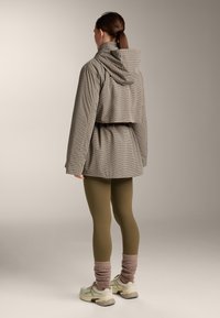 Vrouw met de rug naar de kijker, gekleed in een geruite jas met capuchon, olijfgroene legging, roze losse sokken en beige sneakers, tegen een effen achtergrond.