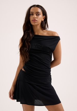 Pikkade pruunide juustega naine, kes kannab suuri rõngaskõrvarõngaid, musta õlgu paljastavat toppi ja sobivat musta seelikut.