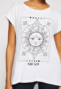 Hvit bomullen t-skjorte med korte ermer, med et svart trykk av en sol design og geometriske mønstre, merket "THE SUN" i svart tekst.