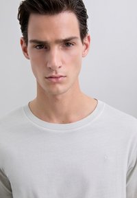 Giovane uomo con capelli scuri che indossa una maglietta bianca a girocollo, che guarda dritto davanti a sé con un'espressione neutra su uno sfondo chiaro e uniforme.