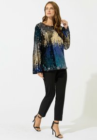 Top con lentejuelas en azul, negro y dorado, con un corte holgado, mangas largas y escote redondeado. Combinado con pantalones ajustados negros y tacones.