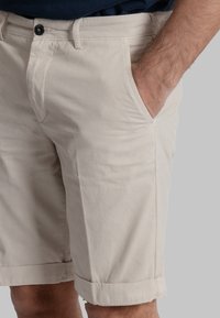 Pantaloni corti beige in cotone con taglio dritto, tasca visibile di lato e orli arrotolati. Presentano una chiusura con bottone nero in vita.