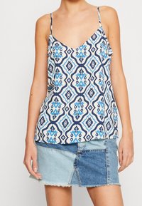 Žena v bezramínkovém vzorovaném topu s modro-bílými geometrickými tvary a dvoubarevných patchworkových džínsových minisukních.