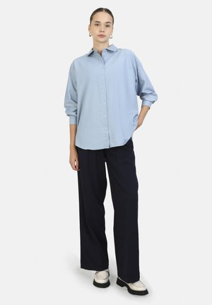 Light blue button-up shirt with long sleeves, smooth fabric, relaxed fit, paired with dark navy wide-leg trousers and cream shoes.