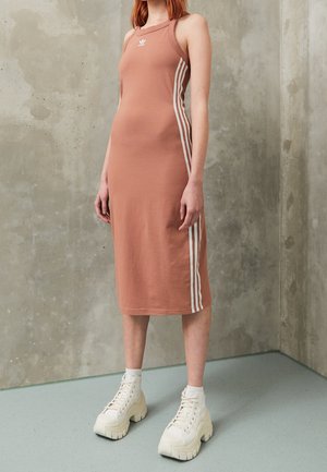 Kobieta ubrana w bezrękawnikową jasnobrązową sukienkę Adidas z białymi paskami po bokach oraz masywne białe sneakersy na platformie, stojąca przy betonowej ścianie.