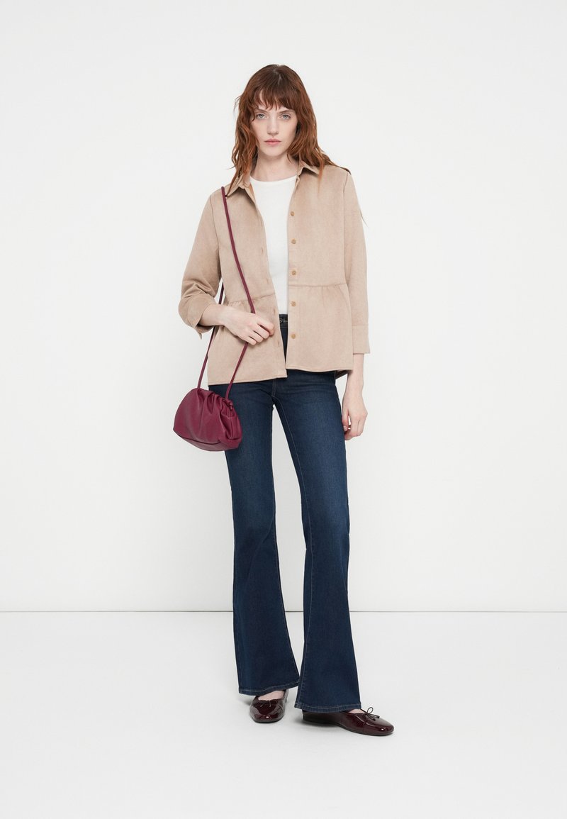 Helle beige Blusenjacke mit Peplum-Design, kombiniert mit dunkelblauen Schlagjeans. Eine kastanienbraune Mini-Handtasche setzt einen kontrastierenden Akzent.