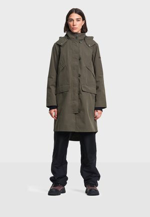 Femme portant un long manteau vert olive à col montant, un pantalon noir et des bottes de randonnée marron, debout devant un fond clair uni.