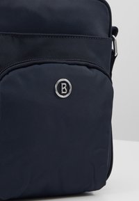 Mała granatowa torebka na ramię z przednią kieszenią na zamek błyskawiczny oraz okrągłym, srebrnym logo na kieszeni.