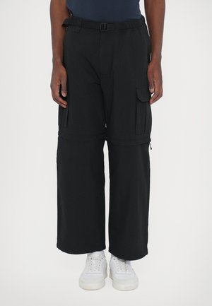 Pantalons cargo noirs à jambes larges, avec de multiples poches latérales, une taille élastique et un détail de boucle ajustable, associés à des baskets blanches.