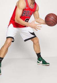 Punainen koripallopaita, jossa lukee "BULLS", valkoiset shortsit mustilla yksityiskohdilla ja vihreät korkeavartiset lenkkarit. Pelaaja pitää koripalloa.