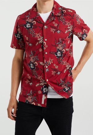 Mann trägt ein rotes kurzärmeliges Hemd mit schwarzem und beigem Blumenmuster über einem weißen Unterhemd und schwarzen Hosen.