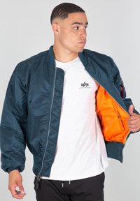 Tmavě modrá bomber bunda se zipem, pružnými manžetami a oranžovou podšívkou. Na hrudi je logo a na boku je kapsa.