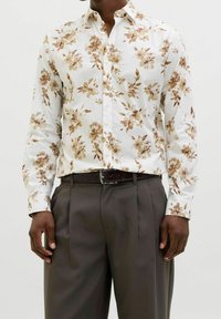 Květinová košile na knoflíky z bílého materiálu s hnědými květinovými vzory. Slim fit s dlouhými rukávy, nošena s tmavě šedými kalhotami a opaskem.