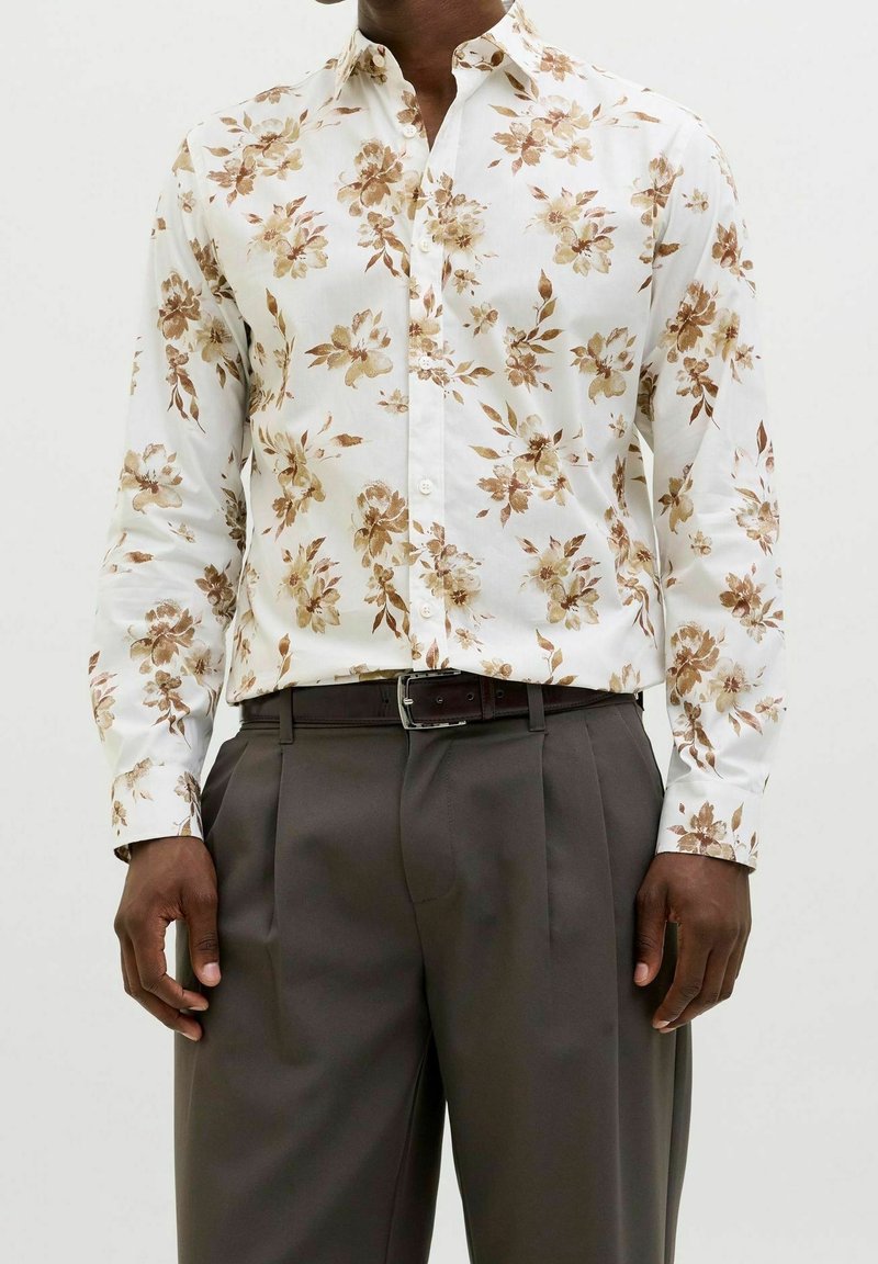 Květinová košile na knoflíky z bílého materiálu s hnědými květinovými vzory. Slim fit s dlouhými rukávy, nošena s tmavě šedými kalhotami a opaskem.