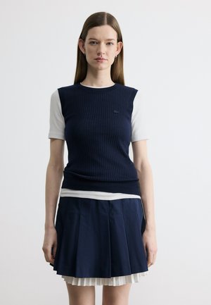 Kobieta o prostych włosach ma na sobie granatowy prążkowany top bez rękawów na białej koszulce z krótkim rękawem oraz granatową plisowaną spódnicę z białą podszewką.
