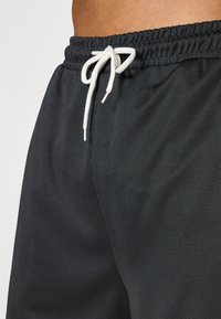 Pantalones cortos deportivos negros con una tela texturizada, cintura elástica y cordón blanco. No se ven bolsillos en la vista recortada.
