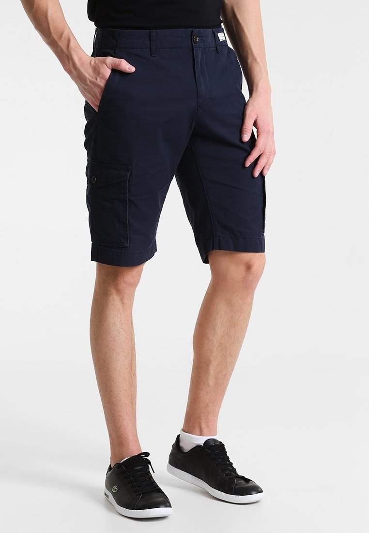Uomo che indossa pantaloni cargo blu navy con tasche con bottoni, sneaker nere e calzini bianchi alla caviglia su uno sfondo chiaro e semplice.