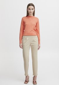 Haut à manches longues orange associé à un pantalon beige ajusté. Les caractéristiques incluent une coupe ajustée, des plis sur le pantalon, et des escarpins ouverts marron.