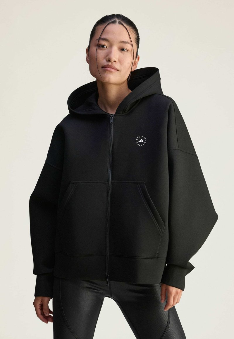 Čierna mikina na zips vyrobená z mäkkej tkaniny s voľným strihom, veľkými prednými vreckami a kruhovým logom na hrudi. Skombinovaná s čiernymi legínami.