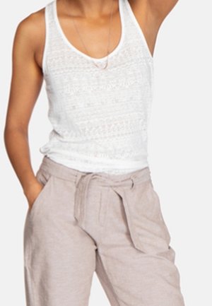 Person trägt ein weißes, ärmelloses Spitzentop und beige, hoch taillierte Hosen mit gebundenem Gürtel, eine Hand in der Tasche, vor einfachem Hintergrund.