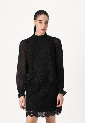 Black lace dress featuring intricate patterns, ruffled high neck, long sheer sleeves, and scalloped hem detailing. Textured floral design.