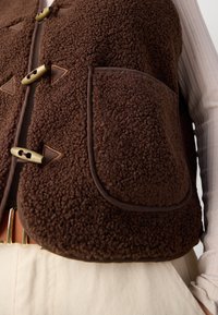 Brown fleece vest featuring a textured surface, brown trim, and two front pockets. Closed with large green wooden buttons.