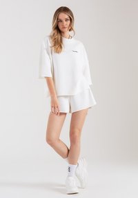 Biała, szeroka bluza z krótkimi rękawami i okrągłym dekoltem, w połączeniu z białymi szortami i białymi trampkami. Prosty, czysty design.