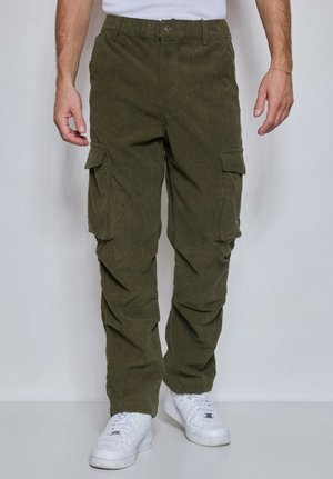 Homme portant un pantalon cargo en velours côtelé vert olive avec poches à rabat et des baskets blanches, debout devant un fond uni.