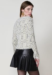 Strukturierter weißer Pullover mit mehrfarbigen Flecken, langen Ärmeln und rundem Ausschnitt, kombiniert mit einem schwarzen, plissierten Lederskirt.