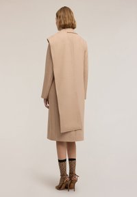 Cappotto di lana beige con un lungo lembo posteriore, silhouette dritta e colletto sartoriale; abbinato a calze nude a fantasia e tacchi beige.