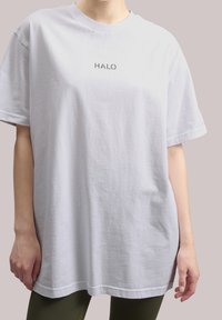 Bílé bavlněné oversized tričko s potiskem "HALO" v šedé barvě uprostřed. Má kulatý výstřih a krátké rukávy, jemnou texturu.