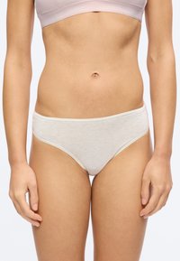 Γυναίκα που φοράει εσώρουχο τύπου σλιπ από βαμβάκι σε ανοιχτό μπεζ χρώμα και ταιριαστό σουτιέν, στέκεται μπροστά σε απλό λευκό φόντο.