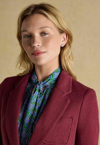 Burgundi põhirelieefiga bleiser rohelise ja sinise mustriga pluusi peal, millel on kaelarihm. Lihtsad hõbedased kõrvarõngad. Neutraalne taust.