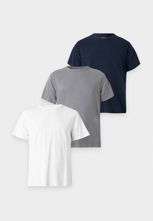 Mand iført en brun kasket, mørkeblå t-shirt, løstsiddende lyseblå jeans og hvide sneakers står med hænderne i lommerne.