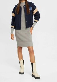 Giacca varsity navy con maniche beige, abito grigio a coste e stivali alti al ginocchio beige e neri con suole testurizzate.