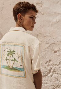 Chemise crème à manches courtes présentant une scène tropicale brodée dans le dos, avec des palmiers, des vagues et des oiseaux dans des couleurs pastel.