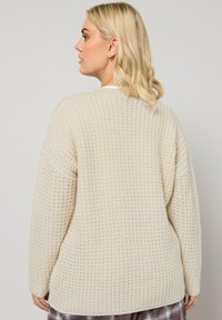 Cardigan en tricot crème avec un motif texturé, coupe décontractée, épaules tombantes et ourlet arrondi, présenté sur un fond neutre.